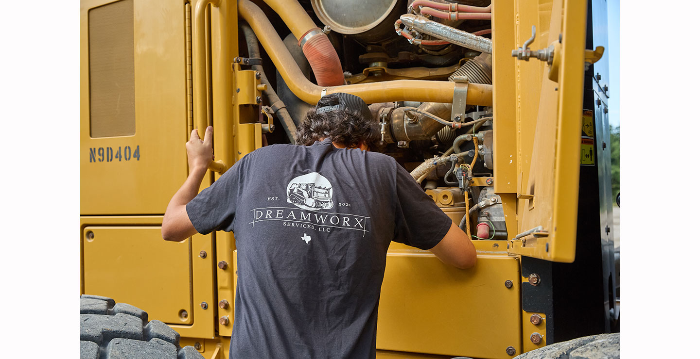 A man, Nick Castillion, wearing a Dreamworx Services T-shirt inspects the open engine compartment of a large yellow construction vehicle.